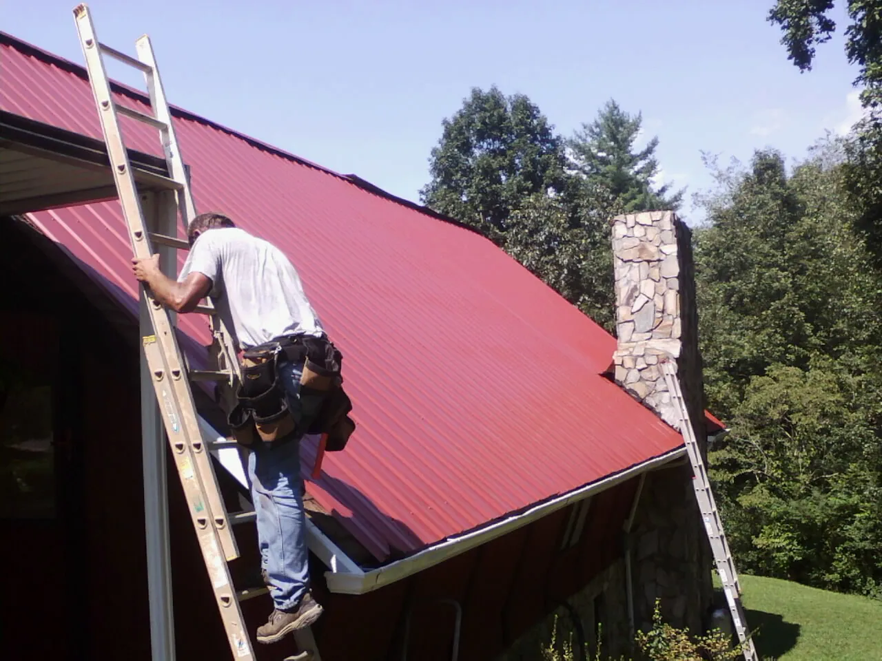 Roofer on a ladder beside a red metal roof and stone chimney — Soffit & Fascia Repair by Roofing Brothers in Wolverton